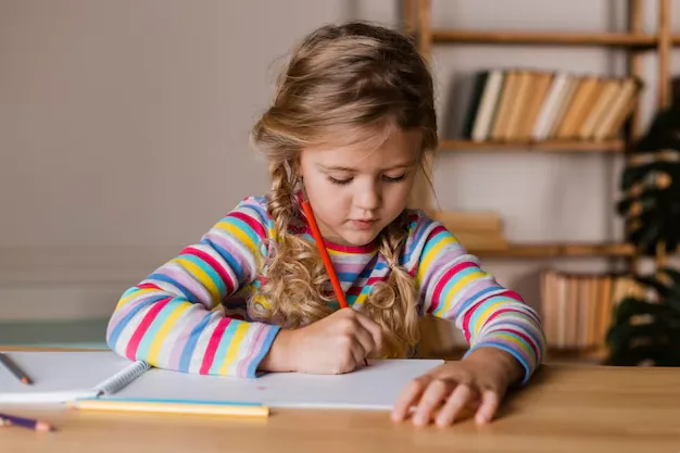 Niña con jersey de rayas coloreando con un lápiz naranja en un escritorio