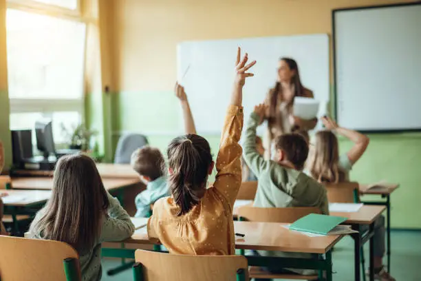 Alumnos levantando la mano en un aula mientras la profesora da clase frente a la pizarra
