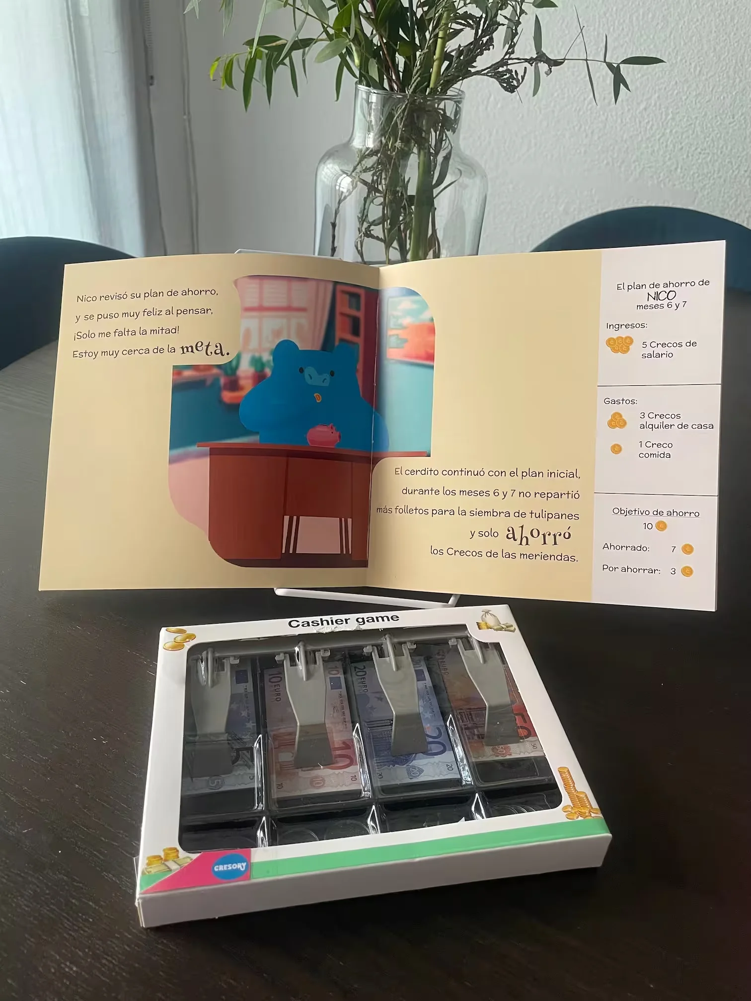 Juego de cajero con billetes de juguete junto a un cuento abierto sobre una mesa